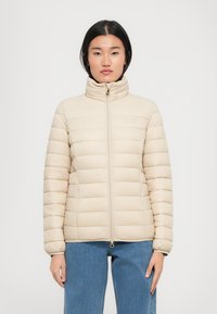 Vrouw in een beige doorgestikte jas met hoge kraag en ritssluiting aan de voorkant, gecombineerd met een blauwe spijkerbroek, staand tegen een effen achtergrond.