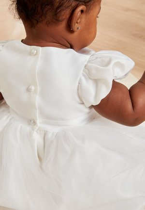Rochie albă cu mâneci bufante, corset din satin și fustă din tul. Are un sistem de închidere cu nasturi din perlă la spate. Material moale cu o textură delicată.