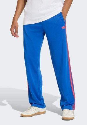 Person trägt blaue Jogginghose mit roten Streifen und Logo an der Seite und weiße Turnschuhe, steht mit einer Hand in der Tasche.