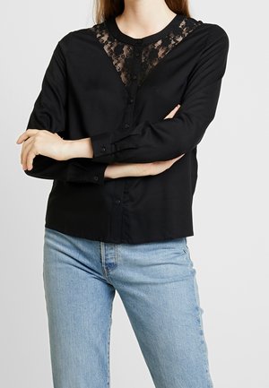 Femme portant un chemisier noir à manches longues avec un détail en dentelle sur la poitrine et un jean bleu clair, bras croisés au niveau de la taille.