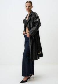 Cappotto trench in pelle nera con ampio colletto, chiusura a bottoni, abbinato a jeans a vita alta in denim blu a zampa d'elefante e tacchi a spillo neri.