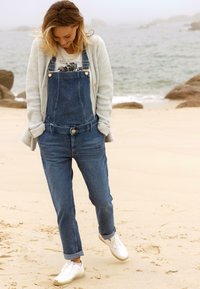 Denim tuinbroeken in een donkere wassing met verstelbare banden, borstzak en een gesloten knoop aan de voorkant, gedragen over een witte graphic tee en gecombineerd met witte sneakers.