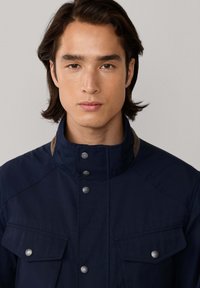 Chaqueta azul marino con cuello alto, botones plateados y dos bolsillos en el pecho. Presenta un acento en contraste de color marrón claro en el cuello. Textura suave.