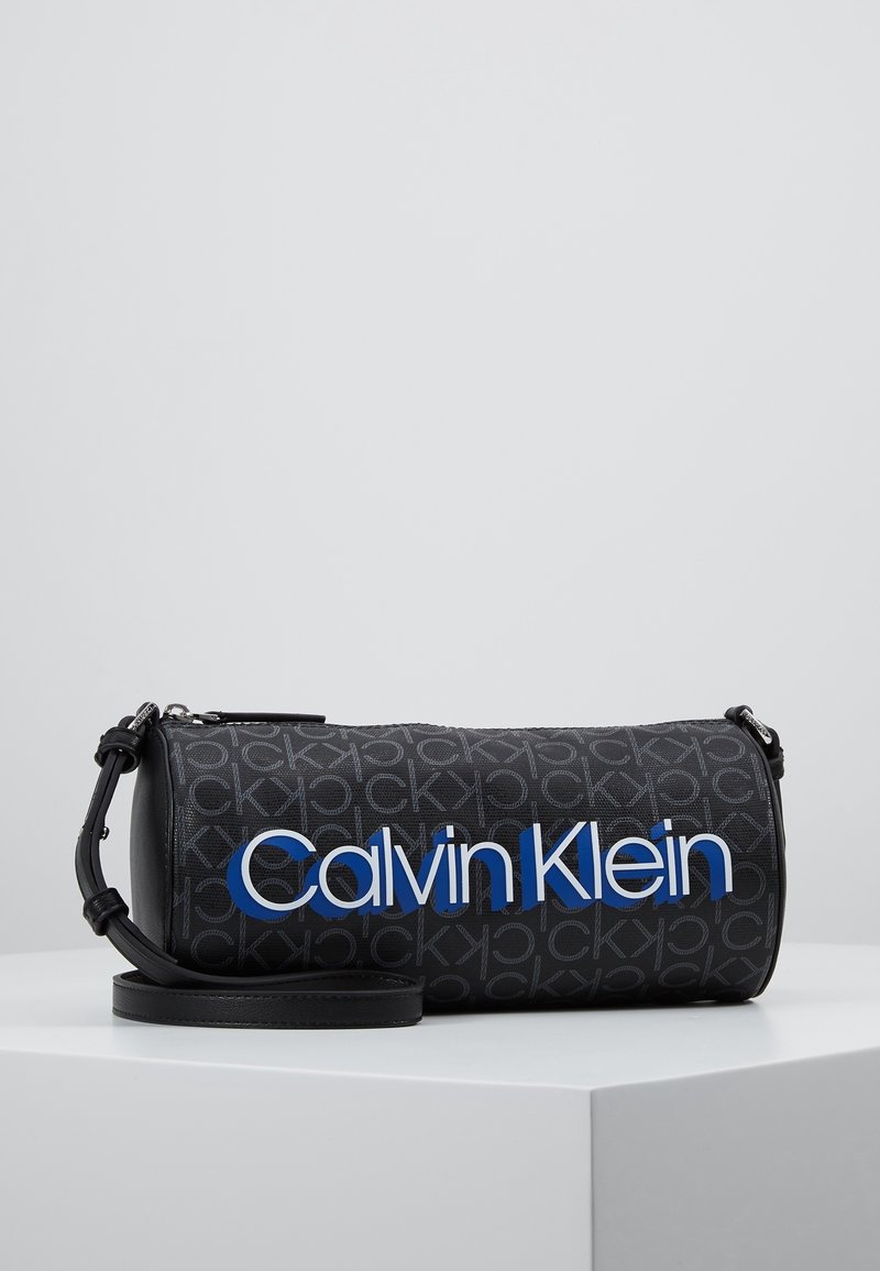 Borsa cilindrica nera con un motivo testurizzato sottile, caratterizzata da un prominente logo blu "Calvin Klein" e una tracolla nera regolabile.