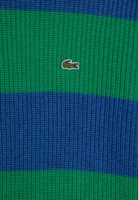 Sweter dzianinowy w niebieskie i zielone poziome paski. Posiada małe wyszywane logo krokodyla. Teksturowana struktura, casualowy design.