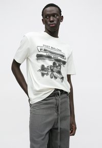 T-shirt de algodão creme com estampa gráfica preta com o texto "POST MALONE" e uma ilustração de um carro, combinado com calças cinzentas com cordão.