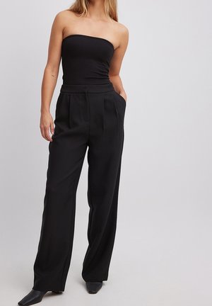 Woman wearing a black strapless top and high-waisted wide-leg black pants with hands in pockets against a plain background.