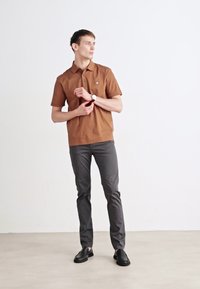 Barna, rövid ujjú pólóing, gallérral, szürke, slim-fit nadrággal és fekete, slip-on cipővel párosítva, egy sima fehér háttér előtt.