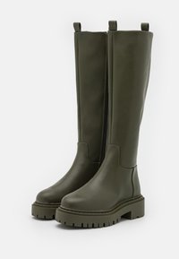 Un par de botas de cuero verde oliva hasta la rodilla con suelas gruesas y cremallera lateral, exhibidas sobre un fondo blanco.