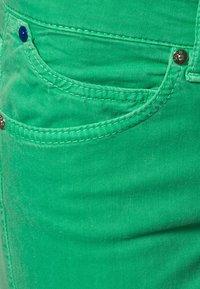 Tela de mezclilla verde en primer plano que presenta un bolsillo frontal con acentos de hardware azul y plateado, costuras blancas en contraste y una textura suave.
