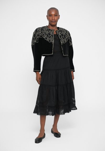 Black velvet cropped jacket with intricate gold embroidery, worn over a tiered black dress. Black flats complete the outfit.