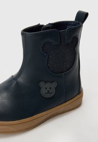 Botín de cuero azul marino que presenta acentos en forma de oso y un panel texturizado. Suela de goma marrón con un acabado suave. Cierre con cremallera.