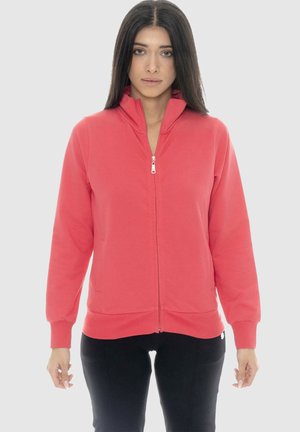 Femme aux longs cheveux foncés portant une veste corail zippée et un pantalon noir, debout devant un fond clair uni.