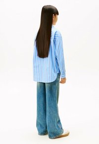 Camicia a righe blu chiaro con maniche lunghe, abbinata a jeans a gamba larga blu e calzature chiare, vista da dietro.