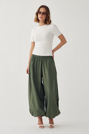 Donna con top bianco a manica corta, pantaloni kaki verde oliva a gamba larga, tacchi bianchi aperti in punta e occhiali da sole sfumati, in piedi davanti a uno sfondo semplice.