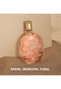 Botella de perfume transparente con líquido de color rosa etiquetada Diesel Fuel for Life 50 ml sobre arena con texto audaz, energizante y floral debajo.