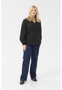 Blouse noire à manches longues avec un col en V et un motif texturé, associée à un jean flare bleu foncé et des chaussures plates noires, sur fond blanc.