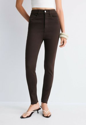 Femme portant un jean skinny marron foncé taille haute, un top court blanc, des bracelets blancs au poignet et des sandales à talons noires avec des lanières blanches.