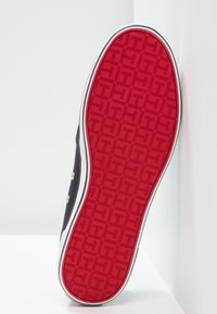 Svart sneaker med en texturerad röd gummisula, vita kanter och metallöglor. Slät ovandel av tyg med sidostickningsdetaljer.