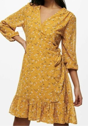 Kobieta ubrana w musztardowo-żółtą, kwiecistą sukienkę kopertową z rękawami do łokcia i falbaną na dole, zawiązaną po prawej stronie w talii.