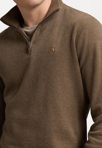 Brauner Pullover mit Viertel-Reißverschluss aus weichem Stoff, mit strukturierter Oberfläche und einem gestickten Logo auf der linken Brust.