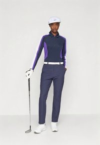 Oseba v mornarsko modrem in vijoličnem golfu, z belo kapo, rokavico in čevlji, ki drži golf palico in stoji pred enostavno ozadjem.