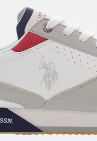Sneaker grigio e bianco con accenti rossi e marina, caratterizzato da perforazioni, suede testurizzato e una sottile grafica polo sul lato.