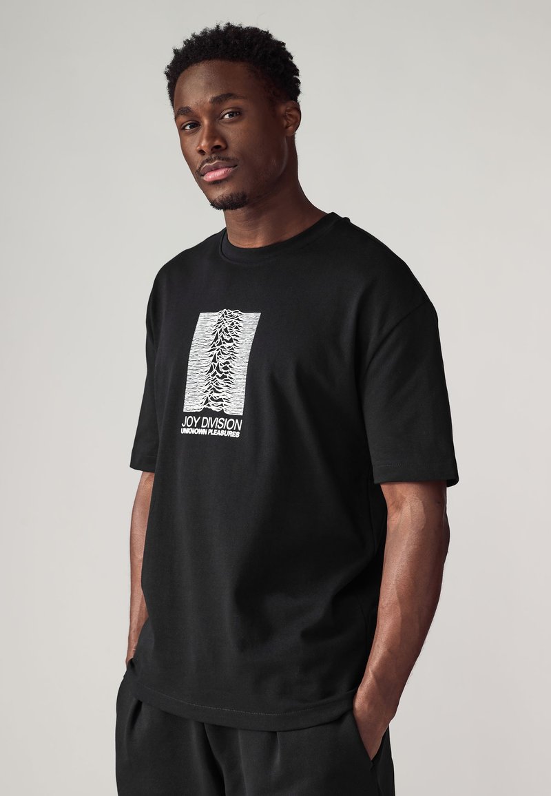 Black cotton t-shirt featuring a white graphic design in the center, displaying wavy lines and the text "JOY DIVISION UNKNOWN PLEASURES."