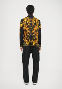 Maglietta nera a maniche lunghe con motivo barocco dorato e dettagli del logo, abbinata a pantaloni cargo neri; scarpe sportive casual completano il look.