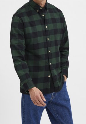 Mand iført grøn og sort ternet skjorte med knapper og lange ærmer og blå jeans, stående med den ene hånd i lommen.
