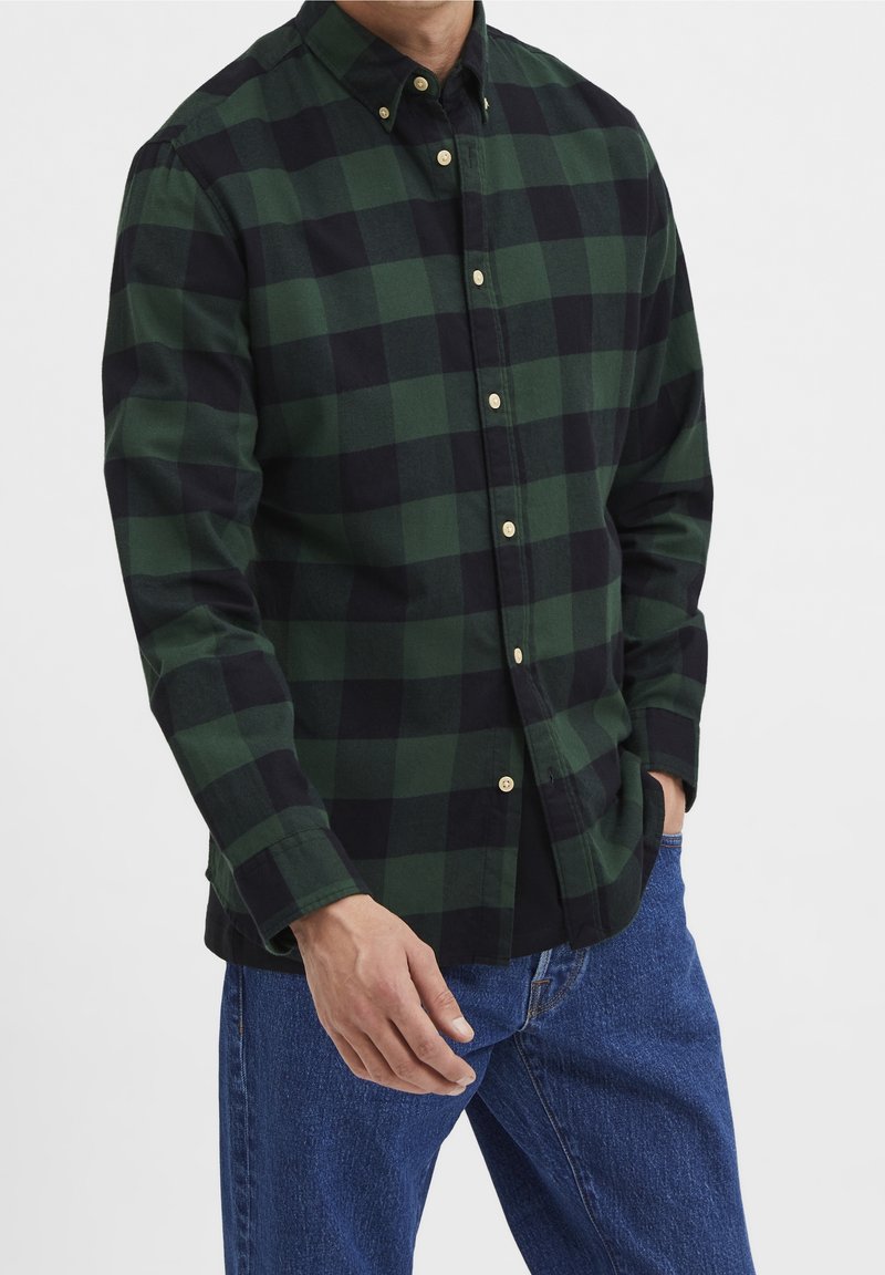 Homme portant une chemise à carreaux verte et noire à manches longues et un jean bleu, debout avec une main dans la poche.
