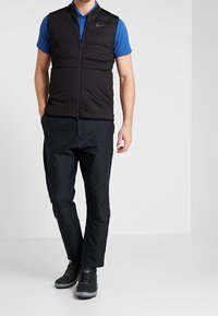 Černá péřová vesta s předním zipem, nošená přes modré polo tričko. Spárovaná s černými kalhotami a černou obuví. Hladká textura, ležérní design.