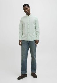 Cămașă verde deschis cu mânecă lungă și guler tip bandă, purtată cu blugi drepți din denim albastru și pantofi închiși la culoare. Design simplu, textură netedă.
