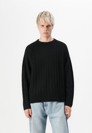 Zwart geribde trui met een ronde hals en verlaagde schouders. Zachte textuur, losse pasvorm en eenvoudig ontwerp. Gecombineerd met lichtblauwe jeans.