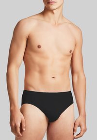 Costume da bagno nero in stile slip, con una texture liscia, vita elastica, design aderente e taglio minimalista, indossato da un modello maschile.