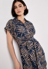 Vestido navy con un suave patrón de hojas de palma en rosa claro, mangas cortas, cuello abotonado y detalle de cintura anudada. Presenta un corte relajado y una textura fluida.