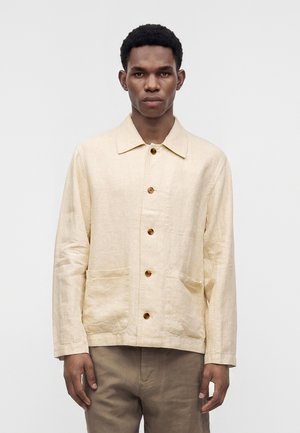 Homme portant une veste beige à boutons avec deux poches avant et un pantalon marron, debout devant un fond clair uni.