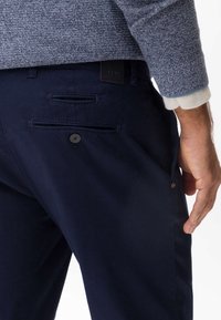 Granatowe spodnie o gładkiej fakturze z ukośnie skośnymi kieszeniami z przodu, dwoma kieszeniami z tyłu zapinanymi na guziki oraz metką marki na pasku.