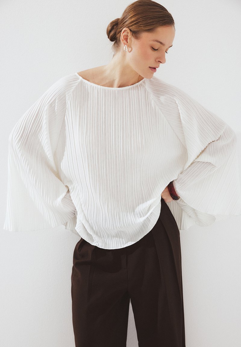 Naine, kellel on juuksed kuklas, kannab volditud valget pluusi laia varrukaga ja tumedaid kõrge vöökohaga pükse, poseerides ühetaolise valge seina taustal.