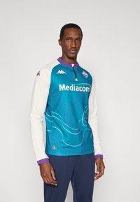 Ilgas rankovių džersis žalsvu atspalviu su baltomis rankovėmis, su bangų raštu, su užtrauktuku ties apykakle ir „Mediacom“ logotipu priekyje. Violetinės detalės.