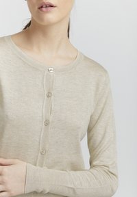 Beige cardigan laget av myk strikket stoff, med rund hals, frontknapper, ribbestrikkede mansjetter og en subtil glans.