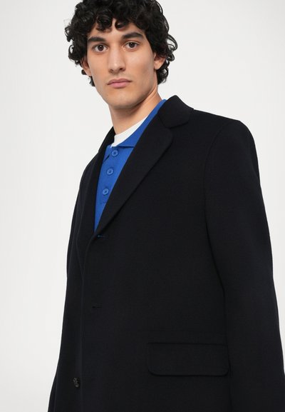 Sacou bleumarin cu o croială ajustată, cu rever înclinat, închidere cu un singur nasture și o buzunar. Purtați peste un tricou polo albastru cu trei nasturi.
