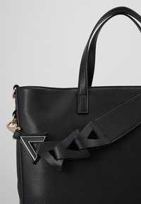 Svart läderhandväska med texturerad yta, utrustad med avtagbart band med geometriska utskärningar och detaljer i guldton.