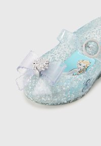 Vaaleansininen läpikuultava kenkä, jossa kimallusta ja koristeellinen violetti rusetti. Kenkässä on hopeinen lumihiutale, ja sisäpuolella on hahmokuva.