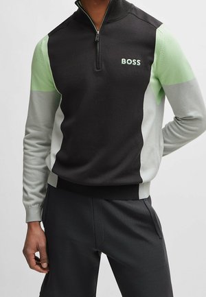 Mężczyzna ubrany w czarno-zielono-szary pulower z przednim zamkiem błyskawicznym i logo "BOSS" na piersi.