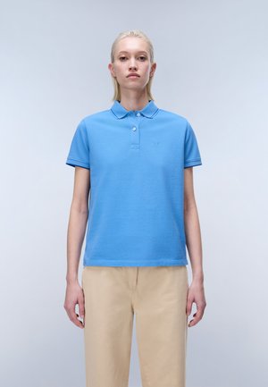 Jeune personne aux cheveux blonds courts portant un polo bleu et un pantalon beige, debout devant un fond gris clair uni.