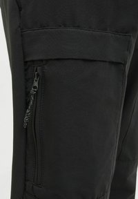 Schwarze Cargo-Hose mit strukturierten Oberflächen, ausgestattet mit einer Reißverschlusstasche an der Seite und einer oberen Klappentasche. Robustes Material, modernes Design.