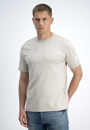Młody mężczyzna z krótkimi włosami, ubrany w jasnobeżowy t-shirt z krótkim rękawem i niebieskie dżinsy, stoi na białym tle, patrząc przed siebie.