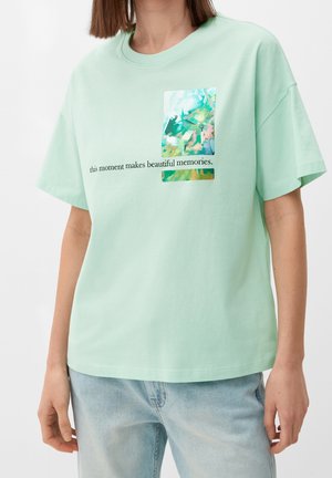 Kvinde iført en lysegrøn t-shirt med abstrakt kunstværk og teksten "dette øjeblik skaber smukke minder," kombineret med lyseblå jeans.