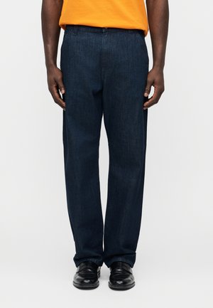 Homme portant un jean droit bleu foncé, des chaussures noires et une chemise orange, debout devant un fond clair uni.